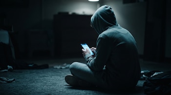 Un joven con sudadera de capucha sentado de espaldas en el suelo de una habitación oscura sostiene un teléfono móvil encendido.