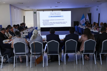 Vista trasera de un grupo de personas sentadas en una mesa de conferencia. Una pantalla grande muestra una presentación sobre el impacto económico de las remesas