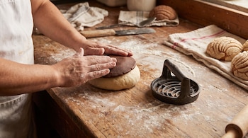 Manos de panadero colocando una cubierta de azúcar de chocolate sobre una bola de masa en una mesa de madera enharinada, con un cortador y conchas al fondo.