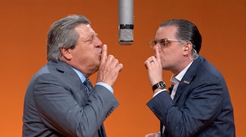 Dos hombres con traje azul y gris colocan el dedo índice sobre los labios frente a un micrófono de estudio suspendido.