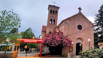 Fachada de iglesia de piedra con campanario y cruz, flanqueada por un árbol de flores rosadas y la entrada a un centro parroquial bajo un cielo nublado