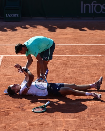 Un tenista de camiseta verde se inclina sobre otro de camiseta blanca, tendido en la cancha de tierra batida. Hay raquetas y marcas de suciedad en la ropa