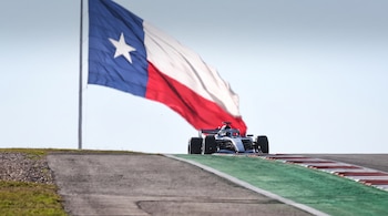 Un coche de carreras monoplaza en una pista asfaltada frente a un mástil con una gran bandera del estado de Texas.