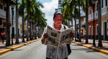 Mujer de rostro enrojecido y ropa sudada sosteniendo un periódico abierto en una calle arbolada con palmeras y edificios al fondo.