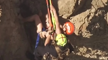 Dos personas con equipo de seguridad, incluyendo cascos amarillo y naranja, manipulan cuerdas o barras mientras realizan una operación en una pared rocosa