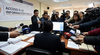 Funcionario de espaldas sentado en escritorio, rodeado por periodistas con micrófonos y cámaras. Un banner al fondo indica 'ACCESO A LA INFORMACIÓN PÚBLICA'.