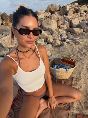 Mujer con gafas de sol, camiseta blanca y bikini oscuro, sentada en la arena con rocas de fondo. Hay un cesto de paja, un sombrero y una cartera
