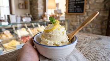 Un tazón blanco con helado de limón, una rodaja de fruta, hojas de menta y una cuchara de madera sostenido sobre una mesa.