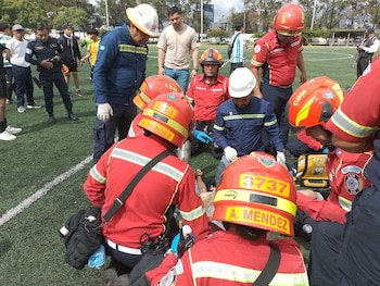 Un hombre con un saco azul y una corbata celeste está sentado en un sofá gris frente a una mesa de vidrio y una ventana