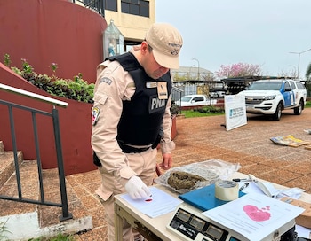Oficial de Prefectura Naval Argentina con uniforme, guantes y chaleco, en una mesa con balanza, documentos, sustancia vegetal. Vehículo utilitario PNA al fondo