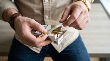 Primer plano de manos sosteniendo una cajetilla de cigarrillos abierta con tabaco y porros de cannabis. La persona usa pulsera de cuero y reloj dorado.