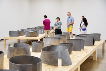 Vista de una galería con paredes blancas, personas y una instalación artística de elementos metálicos oscuros curvos y cilíndricos sobre plataformas de madera