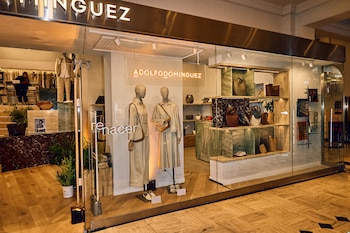 Escaparate de la boutique Adolfo Dominguez con fachada de cristal, exhibiendo dos maniquíes en ropa crema, bolsos y accesorios. Interior iluminado
