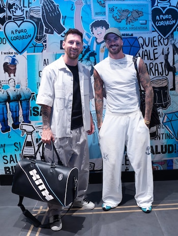 Lionel Messi y Rodrigo De Paul sonríen a cámara, parados frente a una pared decorada con grafitis celestes y negros. Messi lleva un bolso negro