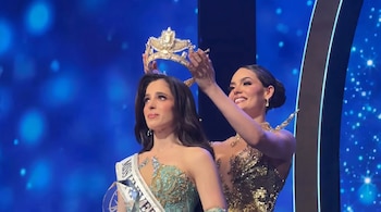 Dos mujeres en el escenario; una sonriente mujer de pelo oscuro coloca una corona dorada sobre la cabeza de otra mujer, que lleva una banda de Miss Universo y un vestido turquesa.