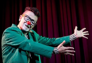 Un hombre disfrazado de payaso con nariz roja, gafas, traje verde y camisa a rayas, extendiendo los brazos, sobre un fondo de cortinas rojas