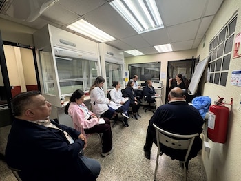 Grupo de profesionales sentados en un laboratorio, algunos con batas blancas, escuchando a una mujer de pie. Hay campanas de extracción y un extintor rojo