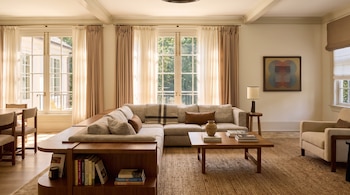 Un sofá en forma de L, una mesa de centro de madera, un sillón y ventanales con cortinas claras en un salón amueblado.