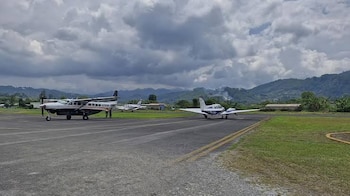 Tres avionetas en una pista de asfalto con césped a los lados, montañas al fondo y nubes densas en el cielo