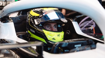 Primer plano de un piloto con casco amarillo fluorescente y negro dentro de un coche de carreras blanco y negro. Se ven logotipos en el casco.