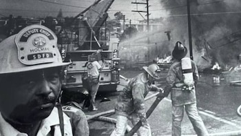 Monocromático. Primer plano de Jefe Adjunto Holman de SDFD, con bigote y casco. Al fondo, bomberos combaten incendio con humo y camión de bomberos en la calle