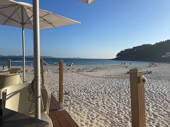 Vista desde un chiringuito con dos sombrillas blancas, sillas, una mesa, postes de madera con cuerdas, arena blanca, mar azul, personas y una colina boscosa
