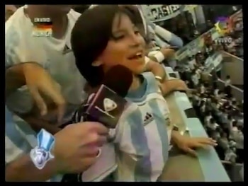 Un joven sonriente con camiseta de fútbol argentina a rayas celestes y blancas habla en un micrófono, rodeado por una multitud en un estadio