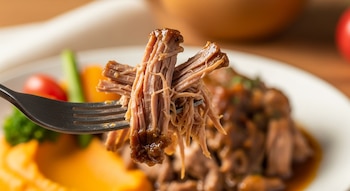 Primer plano de un tenedor sosteniendo carne de cerdo desmenuzada y jugosa. Al fondo, un plato desenfocado con más carne, puré de batatas y vegetales.