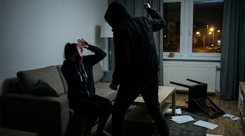 En una sala oscura, una mujer sentada en un sofá se cubre el rostro frente a un hombre encapuchado con el puño levantado. Hay una silla volcada y papeles esparcidos.