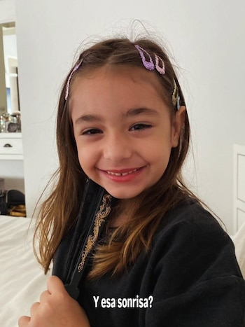 Primer plano de una niña sonriente con pelo castaño, broches brillantes en el cabello y una prenda negra, vista en un fondo claro