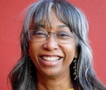 Primer plano de una mujer de piel oscura y cabello gris sonriendo ampliamente, con gafas y pendientes de hojas, sobre un fondo rojo liso