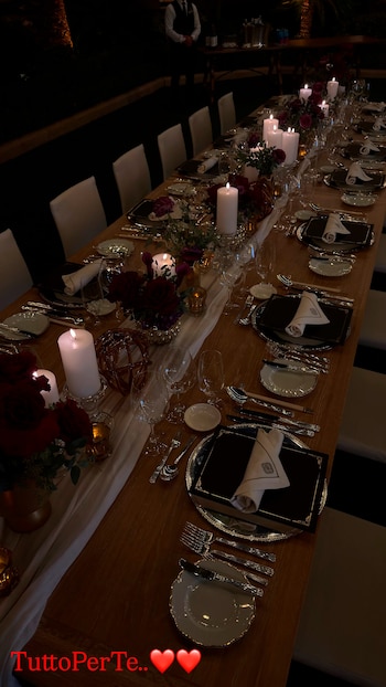 Una mesa de madera alargada vestida con un camino blanco, velas encendidas, arreglos de rosas rojas, vajilla, cubiertos y copas de cristal