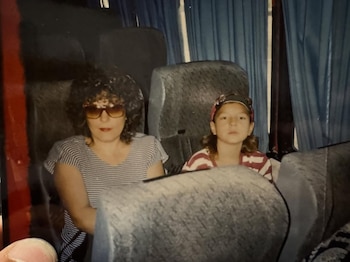 Una mujer con pelo rizado y gafas de sol, junto a un niño con gorra, sentados en asientos de autobús con cortinas azules en el fondo
