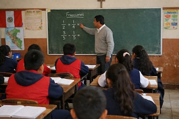 Un profesor frente a una pizarra con problemas de fracciones, y varios alumnos sentados de espaldas en sus pupitres. Se ven libros, una bandera de Perú y un mapa.