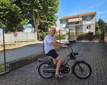 Hombre calvo con camiseta blanca y pantalones cortos negros sonriendo mientras está sentado en un ciclomotor Garelli gris en un camino adoquinado