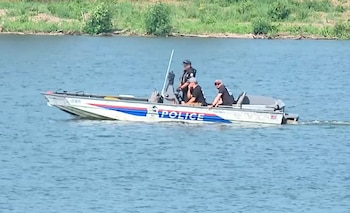 Un barco policial con tres hombres a bordo navega sobre el agua. Se observa una franja de vegetación en la orilla del río al fondo
