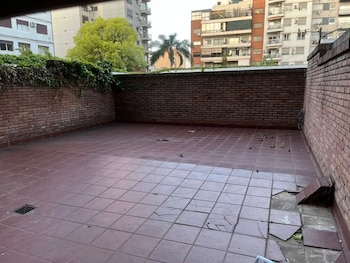 Un patio exterior descuidado con baldosas de terracota partidas, paredes de ladrillo vistas, follaje y edificios residenciales en el fondo