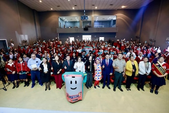 Grupo grande de estudiantes uniformados y adultos posa en un auditorio con un muñeco de la Línea ANNA 1810; los estudiantes sostienen tarjetas azules