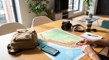 Manos extendidas sobre un mapa impreso del Perú en una mesa de madera con una mochila beige, una cámara, un teléfono móvil y una libreta.