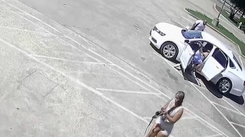 Vista aérea de una mujer con cabello claro sosteniendo un rifle en un estacionamiento, junto a un sedán blanco con las puertas abiertas