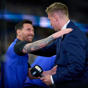Lionel Messi sonríe mientras coloca la mano sobre el hombro de un hombre con traje azul que sostiene un micrófono