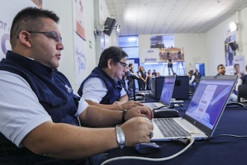 Dos hombres con chalecos de la ONPE operan laptops en una mesa. Al fondo, otras personas y equipos de grabación en un centro de procesamiento electoral