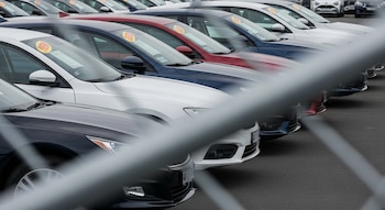 Vista en diagonal de varios autos nuevos de diferentes colores estacionados en una fila, con etiquetas de precio, observados a través de una valla metálica gris borrosa en primer plano.