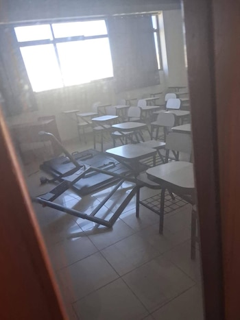 Un aula universitaria con pupitres, una pizarra o pantalla de proyección caída en el suelo y una atmósfera polvorienta, vista a través de una abertura
