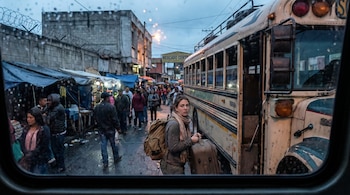 Vista desde dentro de un vehículo de una mujer con mochila y bufanda bajando de un autobús en una terminal de Guatemala, sujetando una maleta en una calle mojada con puestos y gente.