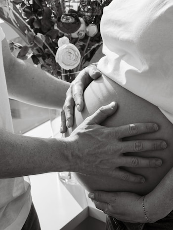 Fotografía en blanco y negro de un vientre de embarazada. Manos de dos personas lo tocan. Un ramo de flores aparece difuso en el fondo
