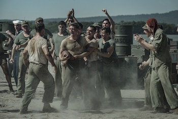 Varios hombres vestidos con uniformes militares celebran en un terreno de tierra junto a un joven que sostiene una pelota de fútbol americano