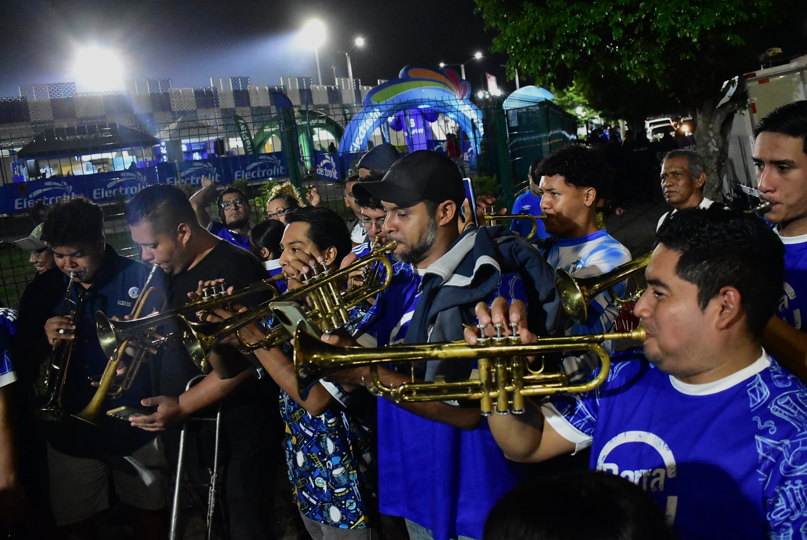 Un grupo de músicos vestidos con camisetas azules toca instrumentos de viento de metal cerca de un recinto iluminado.