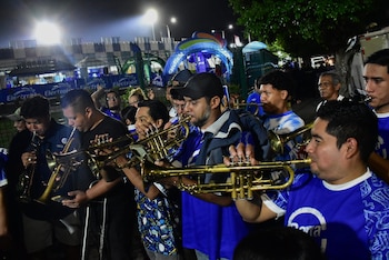 Un grupo de hombres sostiene y toca trompetas y trombones de metal dorado durante la noche frente a una valla