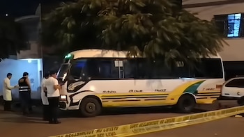 Un minibús blanco estacionado de noche en una calle con personas al lado y una cinta de seguridad amarilla en primer plano